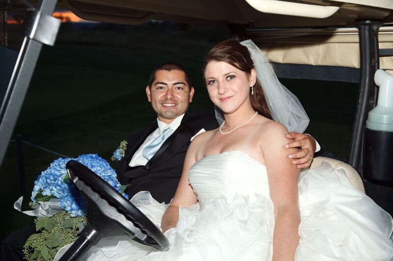 Newlyweds in a golf cart. Man in suit, woman in a white wedding dress and veil, holding bouquet.