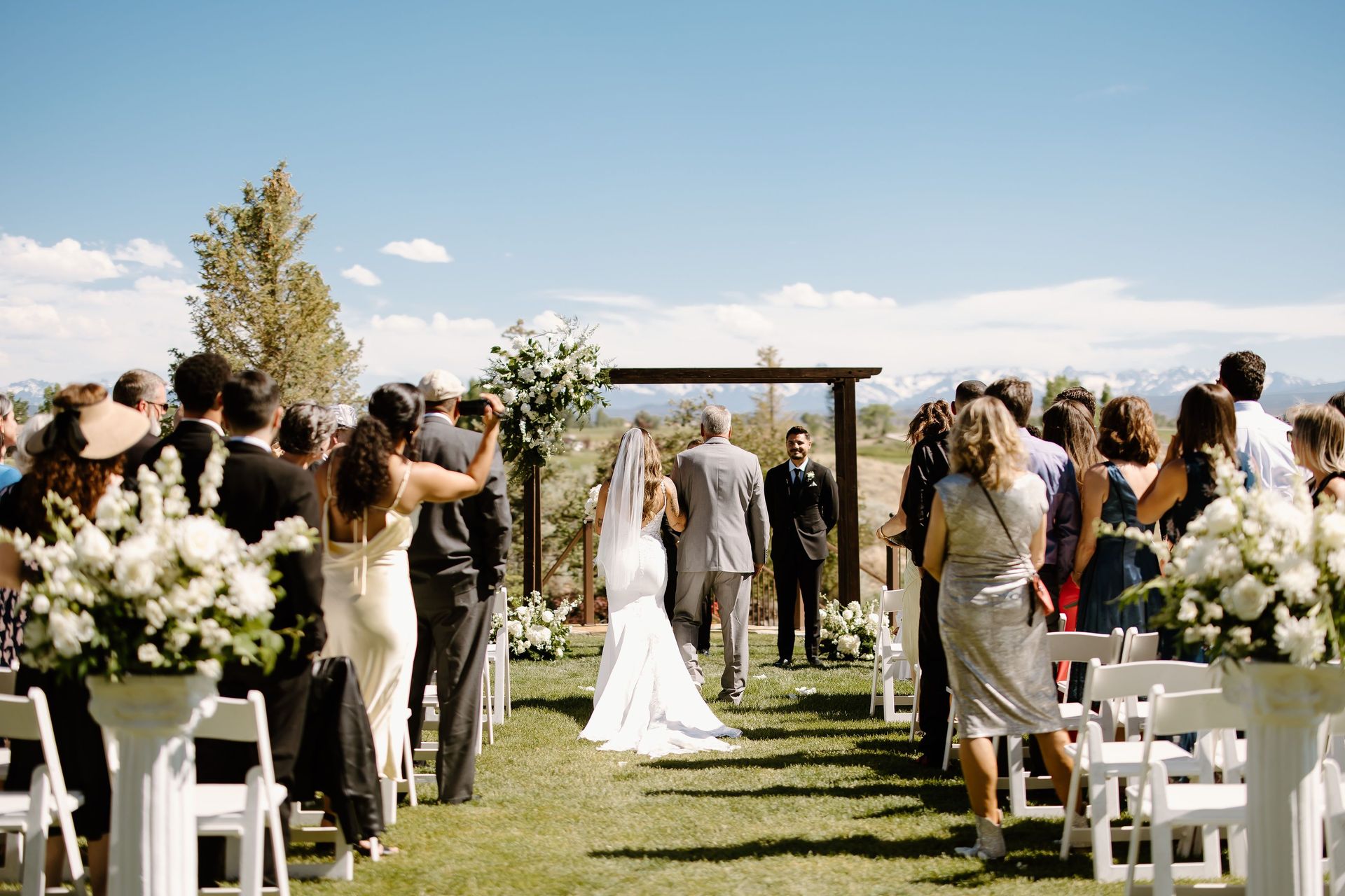 Bride escorted down aisle at outdoor wedding ceremony. Guests, floral arrangements, arch, and blue sky visible.