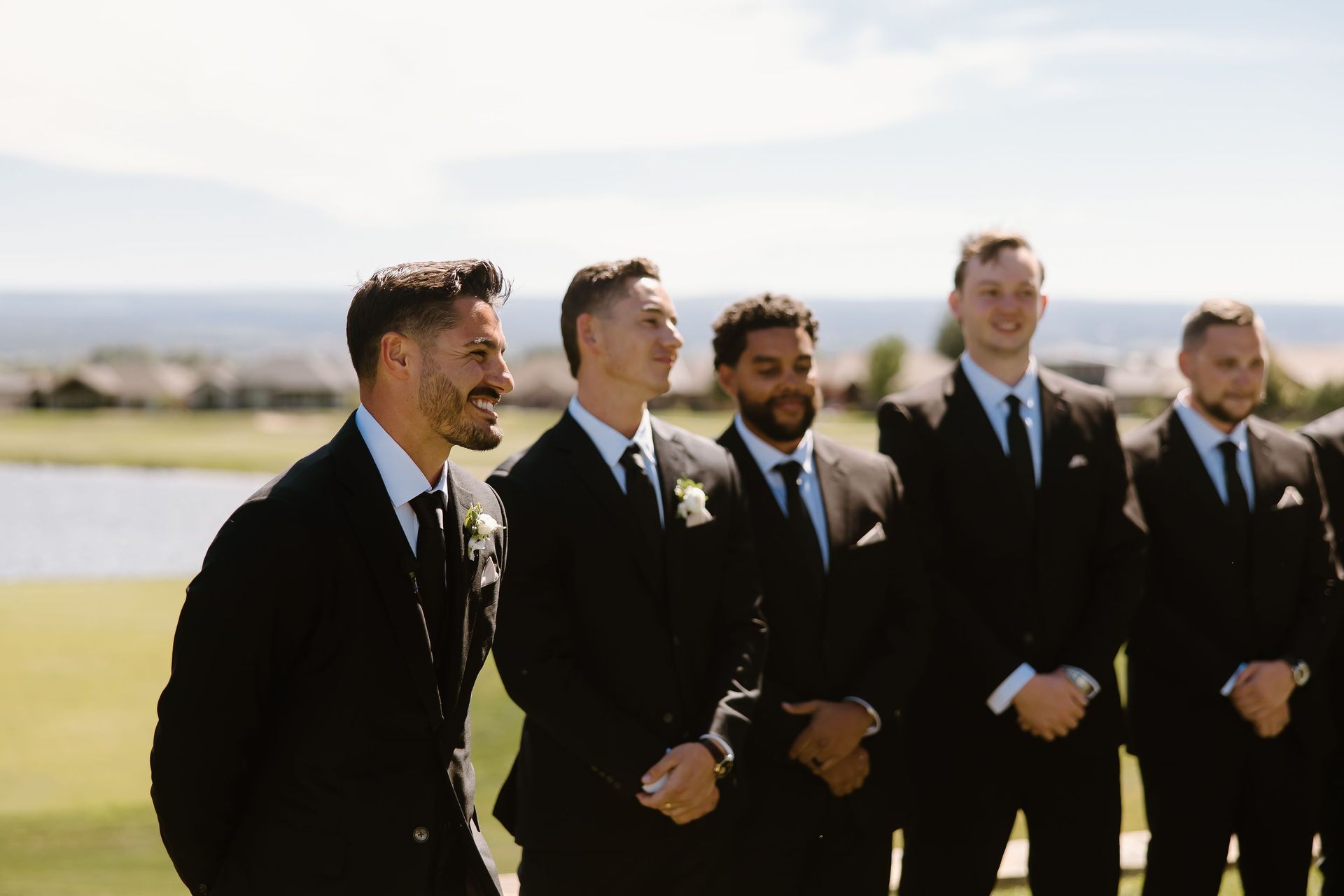 Men in black suits stand outdoors, likely at a wedding, smiling and looking off-camera near a golf course.