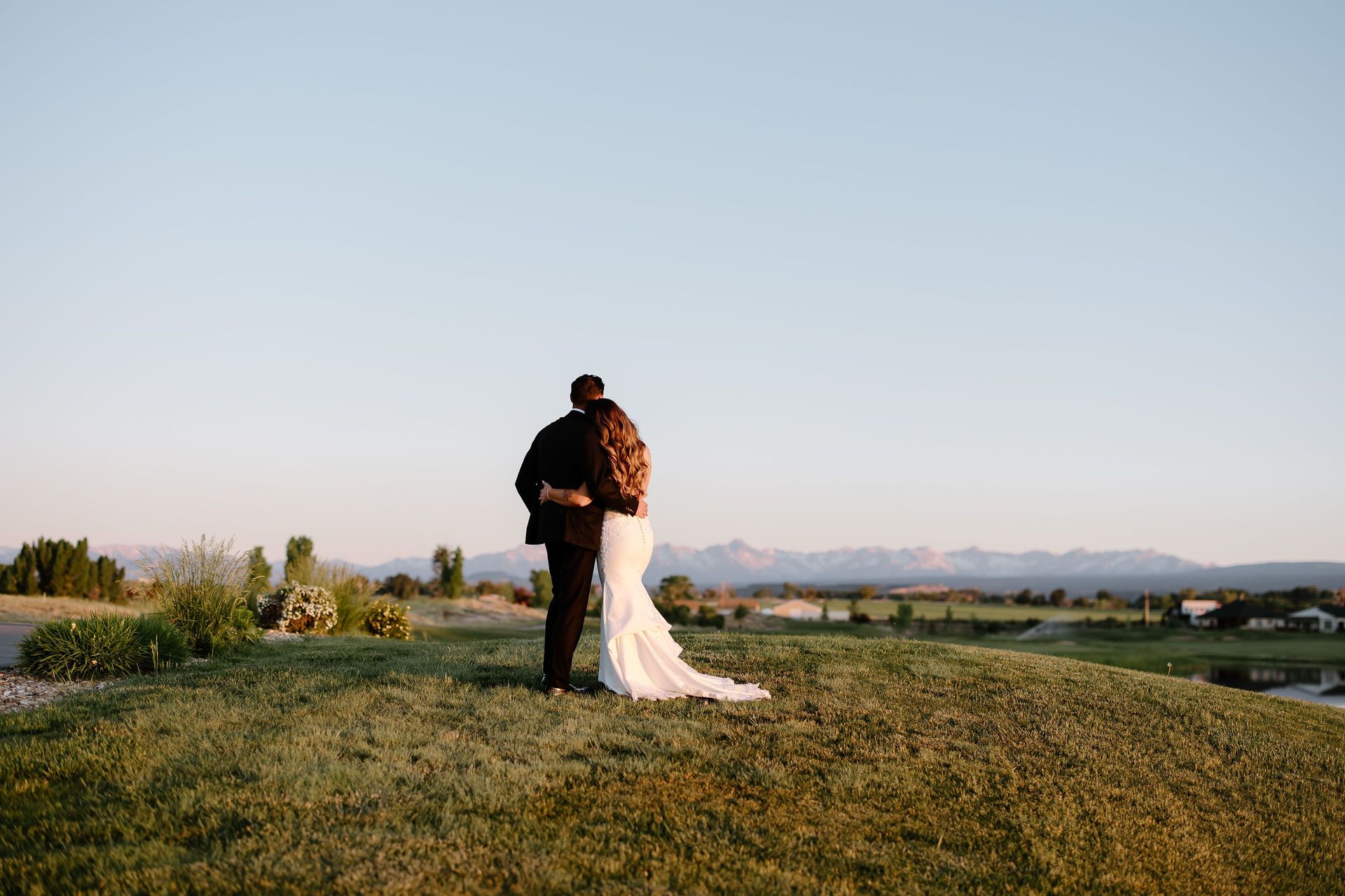 Couple embracing on a grassy hill overlooking a valley, mountains in the distance; golden hour lighting.
