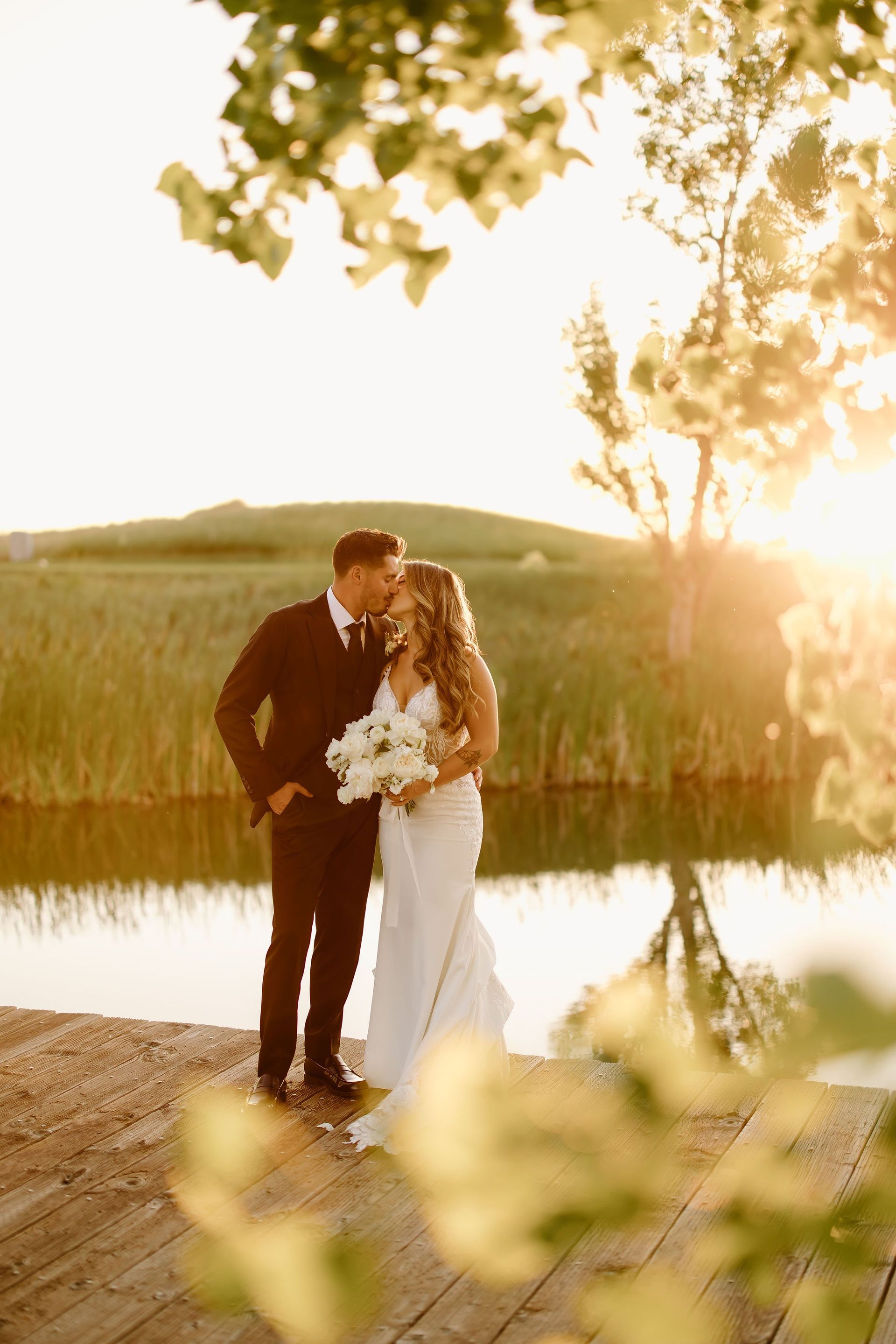 Couple kissing on a wooden dock by a lake at sunset. The bride wears a white dress and holds flowers.