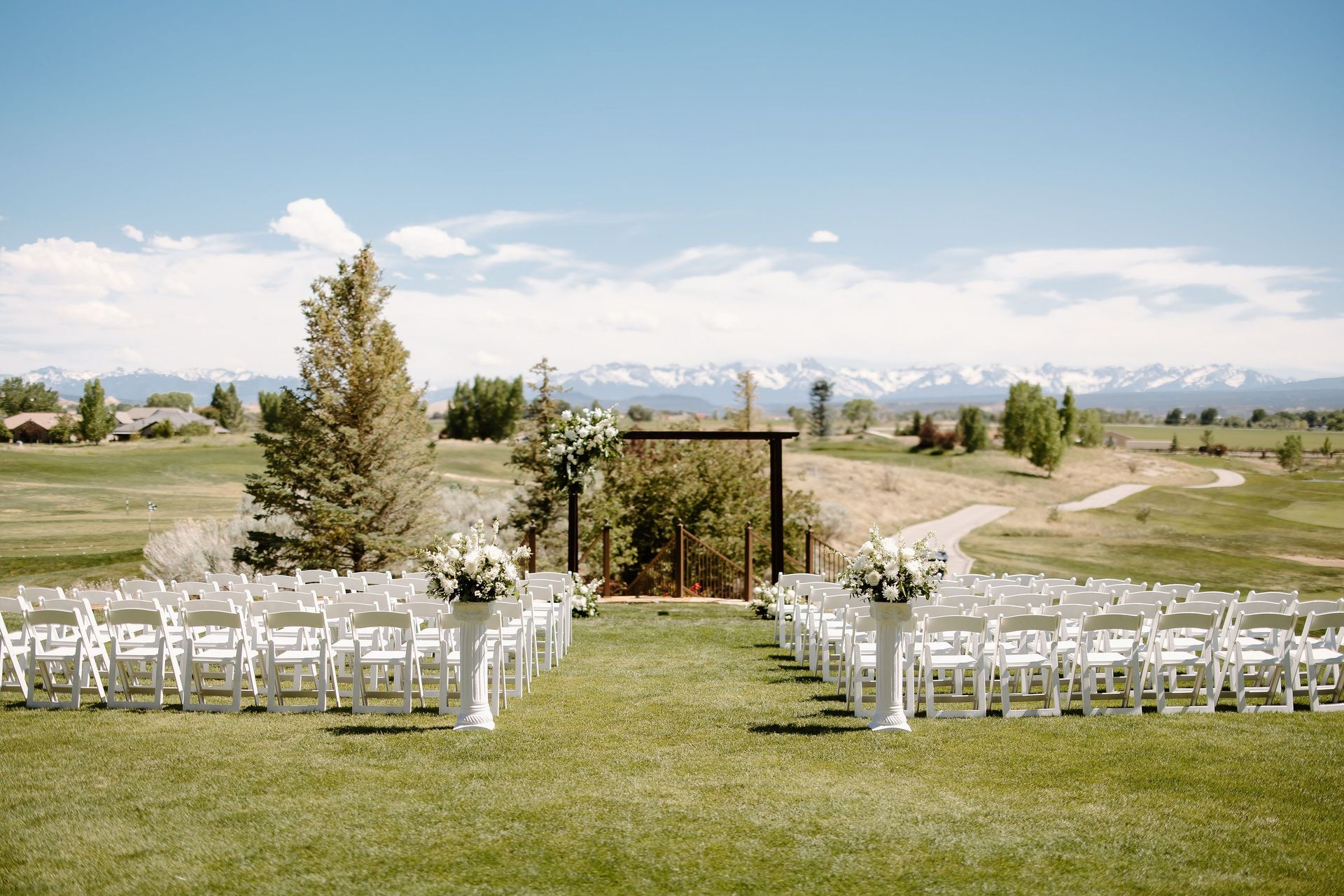 Wedding ceremony setup on a grassy hill with white chairs, floral arrangements, and a mountain backdrop.
