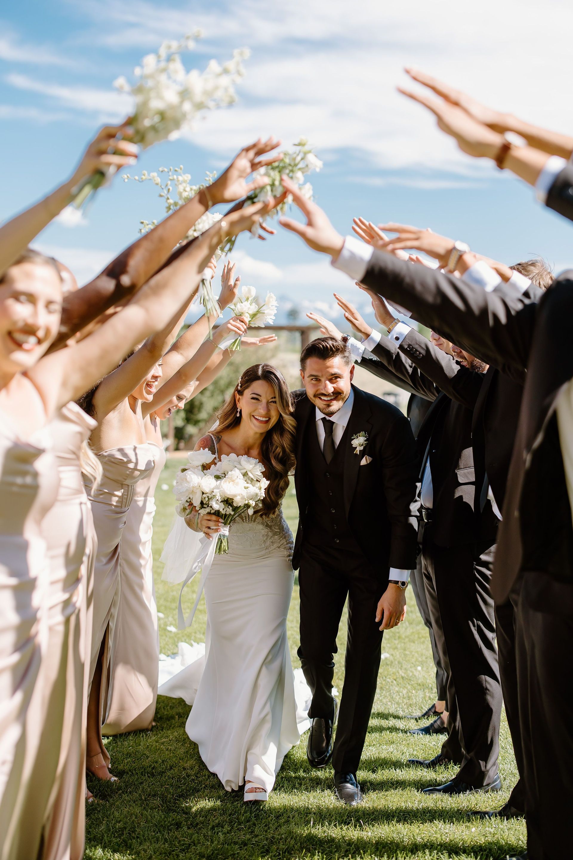 Wedding couple walks under an arch of raised arms, smiling. Outdoor, sunny setting.