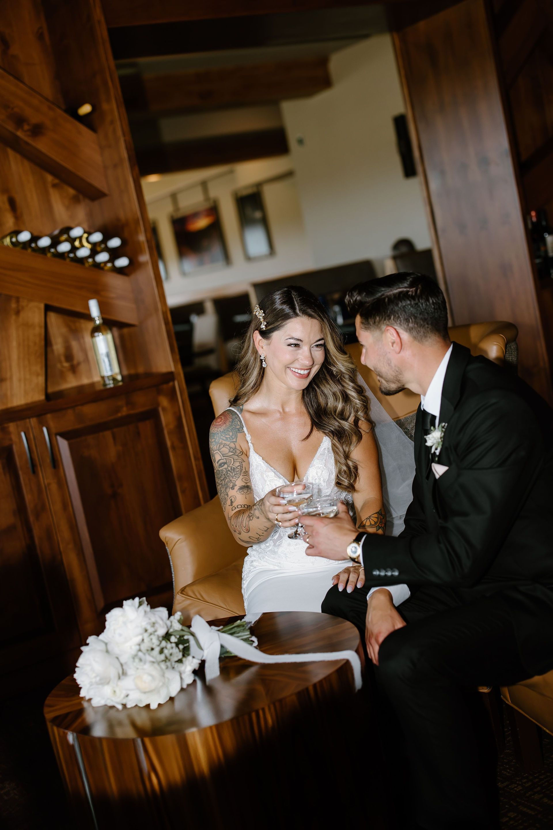 Wedding couple, bride in white dress with arm tattoos, groom in black suit, smiling. Indoor setting with wood accents.