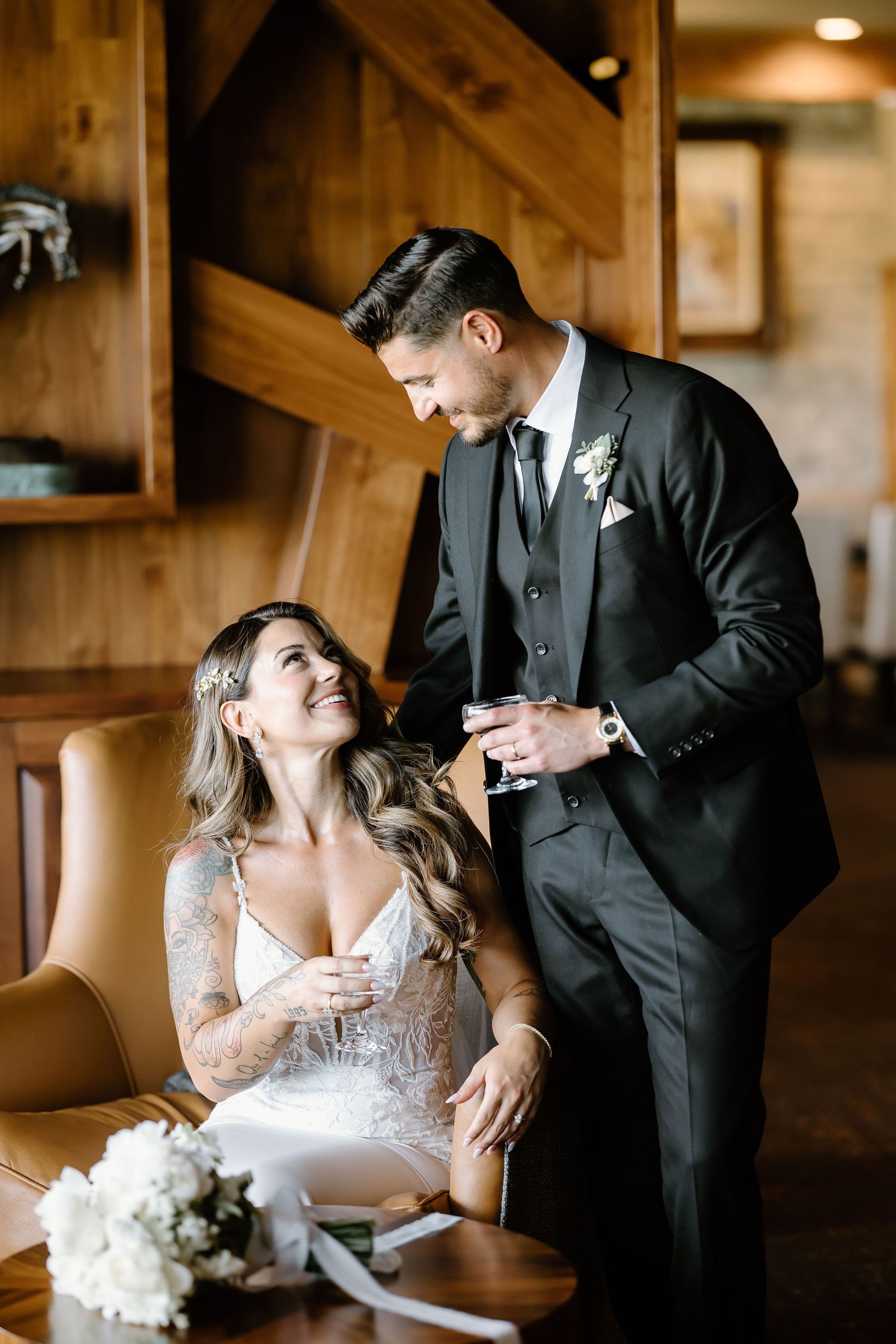 Bride and groom gazing at each other. She sits in a chair; he stands holding a drink. Wooden interior.