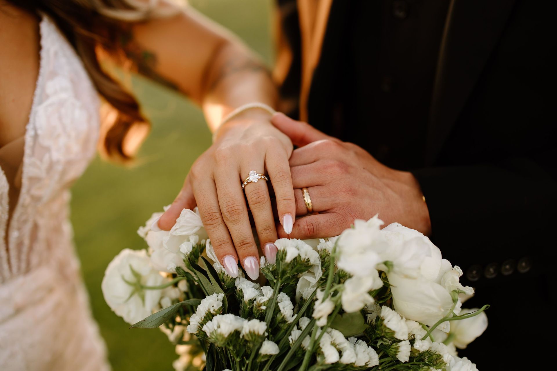 Close-up of hands with wedding rings, holding a bouquet of white flowers, in a sunny outdoor setting.