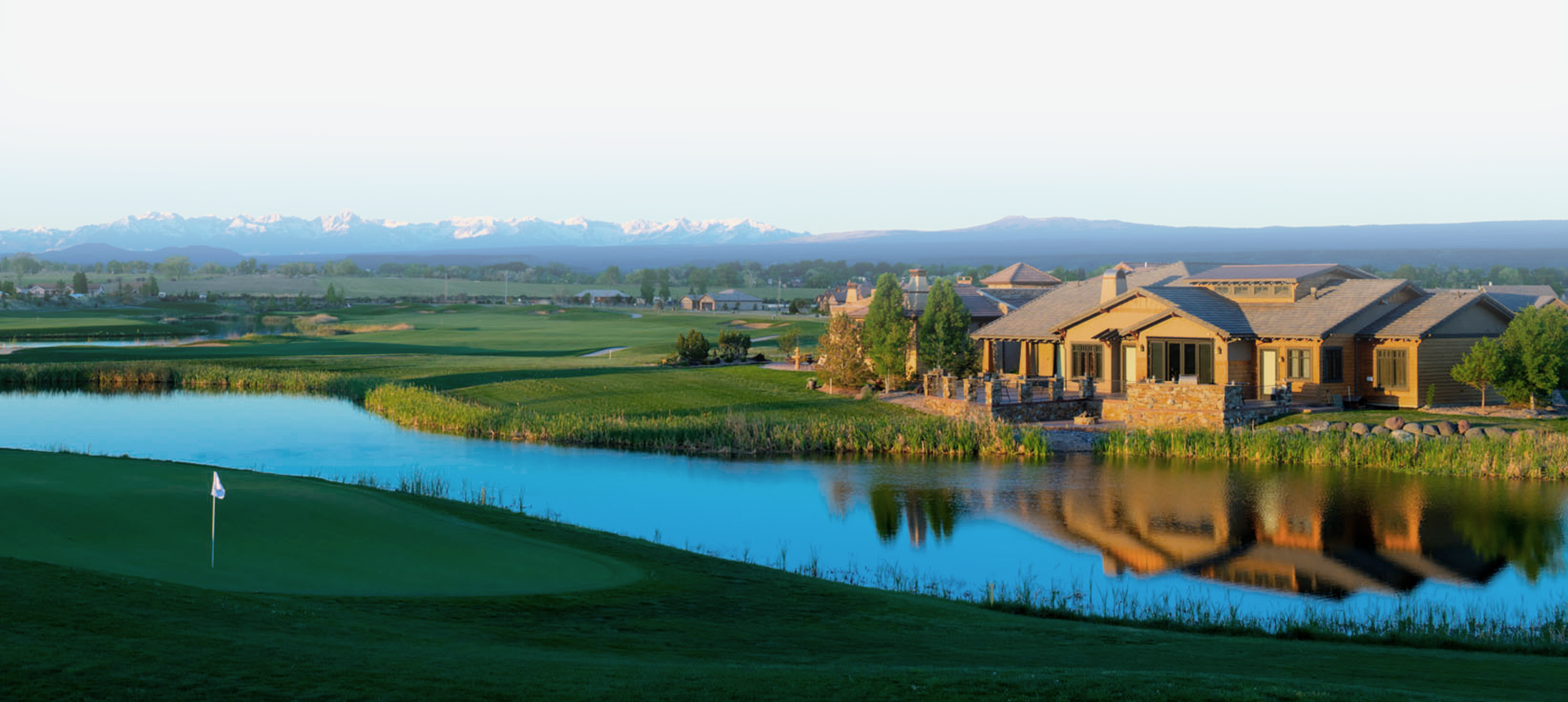 Golf course with water feature, clubhouse, and green fairway under a light sky.