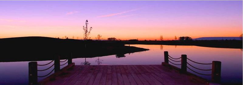 Wooden dock extends over a calm lake reflecting the purple and orange sky at sunset.