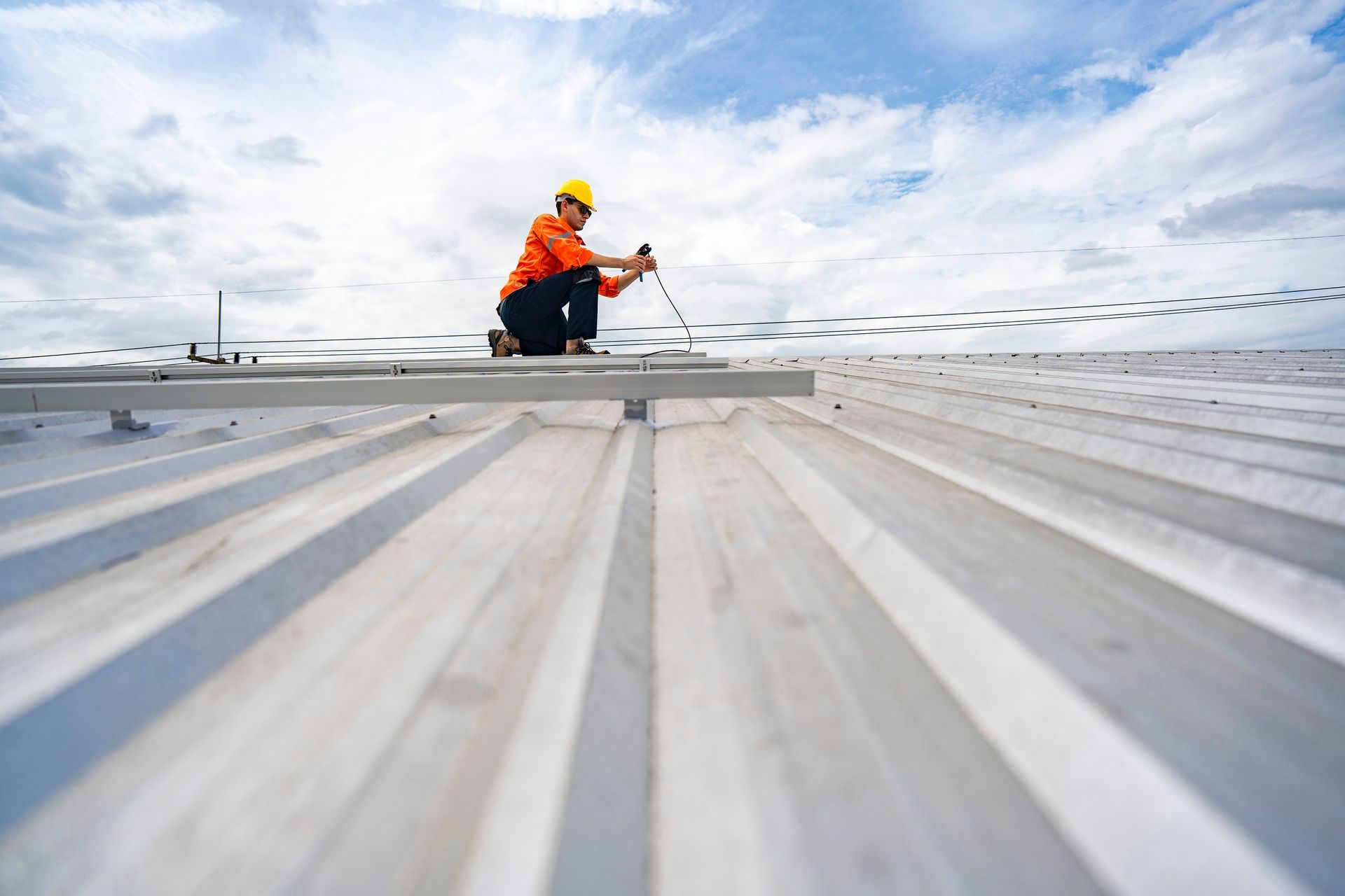 A worker is installing metal roofing panels using safety gear under a cloudy sky