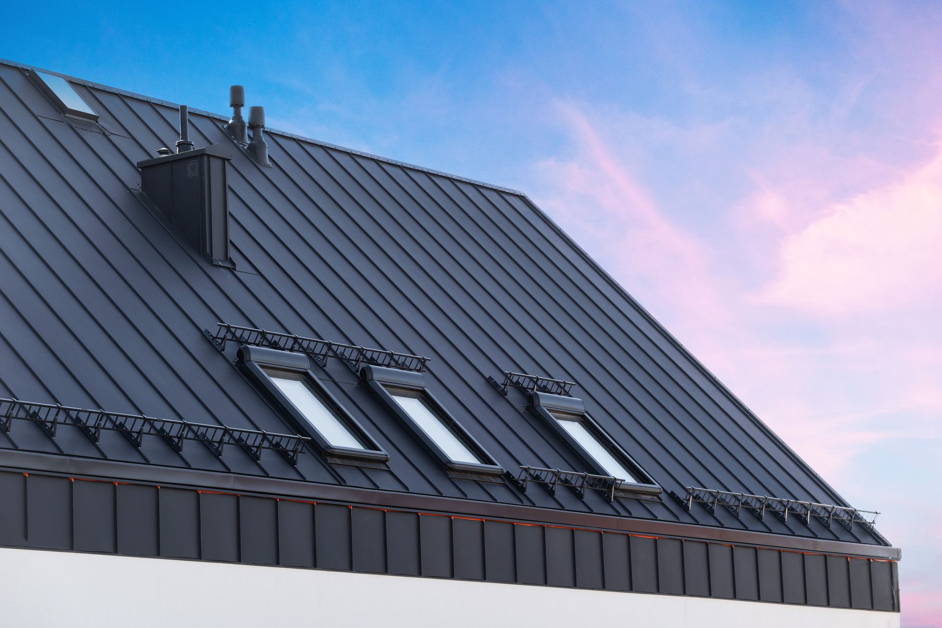 Black metal roof with skylights and chimney against a blue and pink sky.