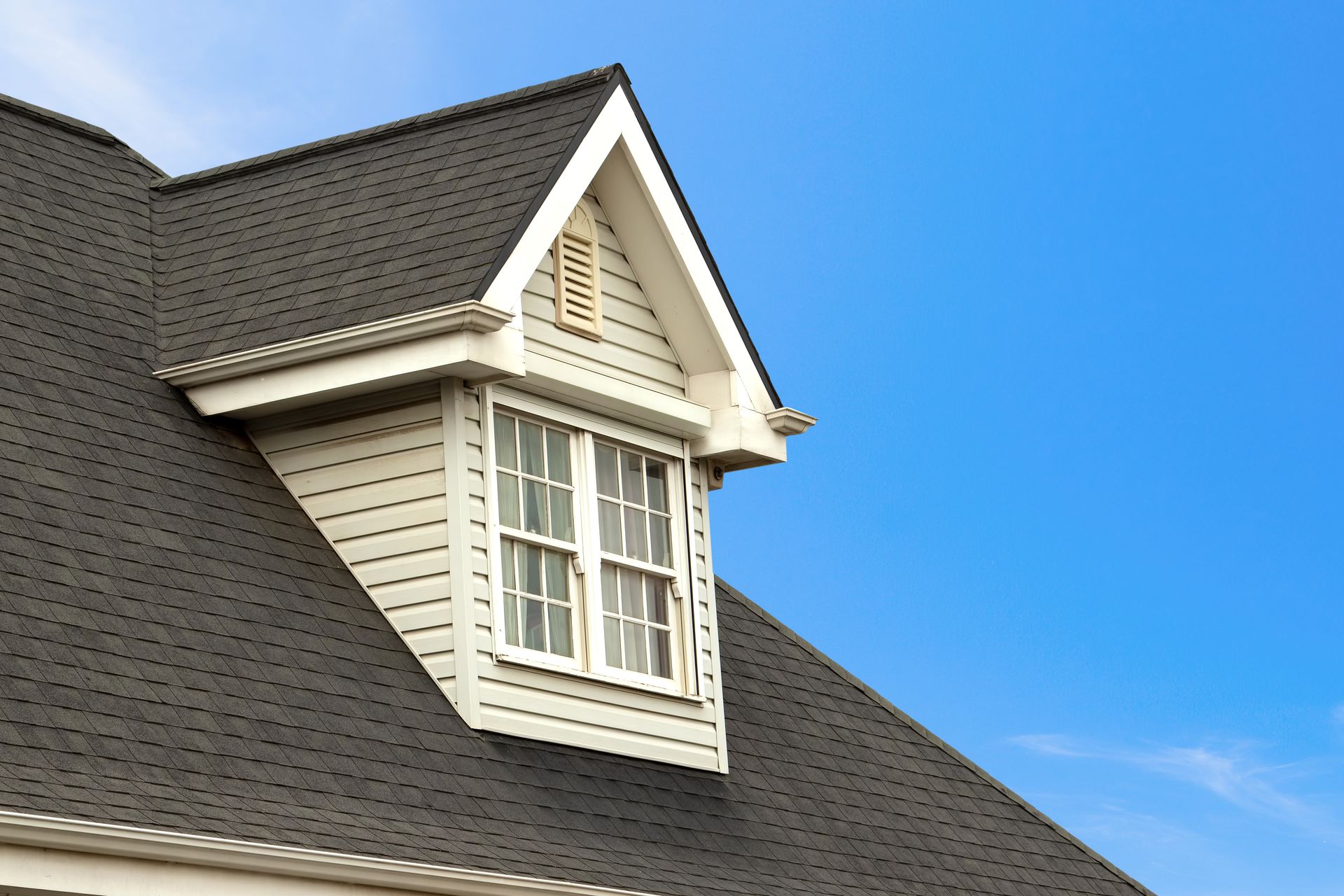 Close-up of a dormer window on a roof, white siding and trim, dark gray shingles, against a blue sky.