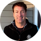 Man smiling, wearing a black jacket, standing outdoors in front of a building.