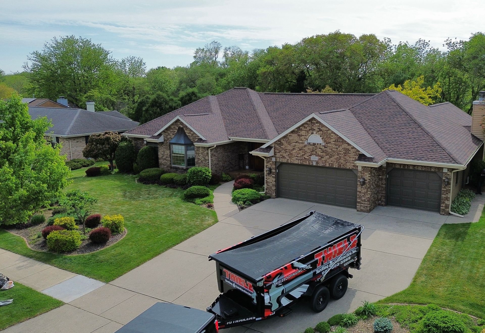 House with brown roof, stone facade, and attached garage. A trailer with a red and black graphic is in the driveway.