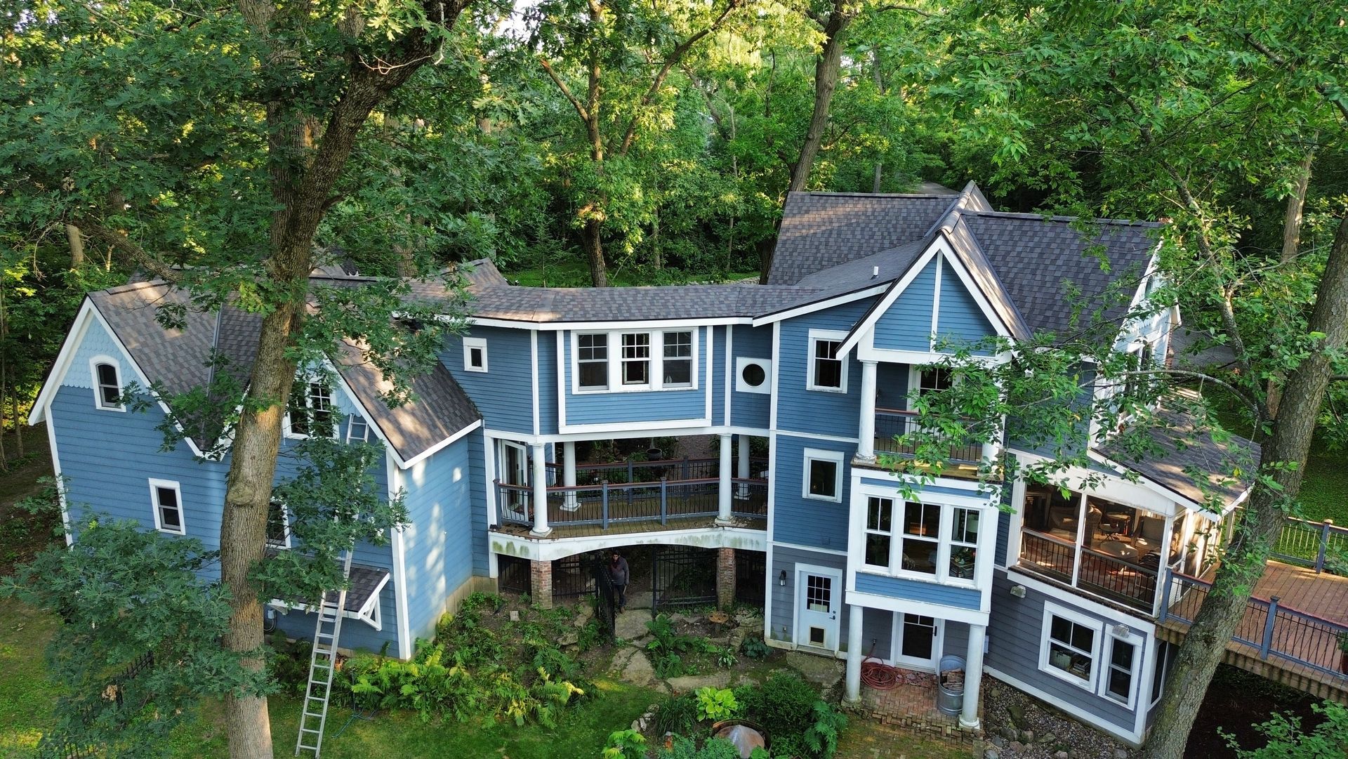 Blue multi-story house surrounded by trees. White trim, multiple balconies, dark roof.