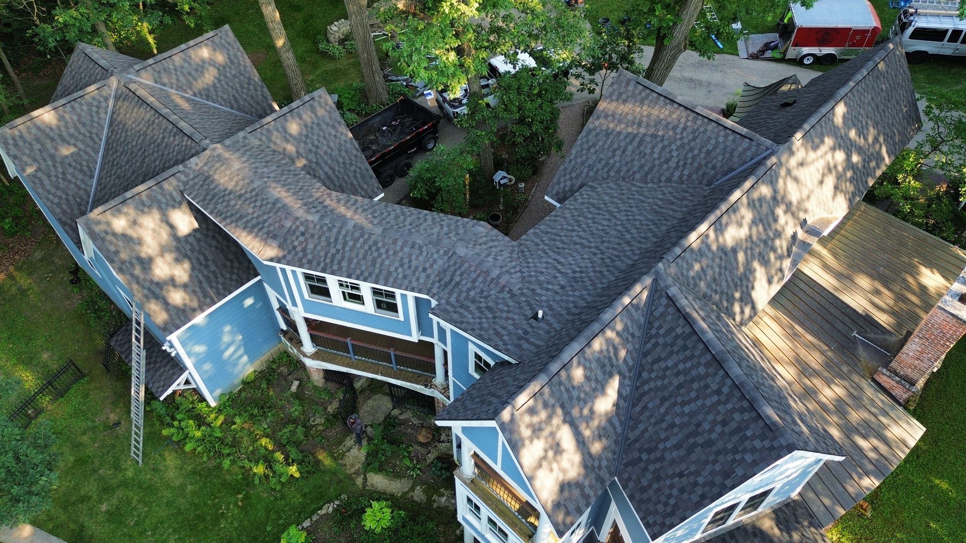 Aerial view of a blue house with multiple roof sections. Green yard and trees surround it.