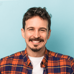 Man with dark hair and mustache smiling, wearing orange plaid shirt, against blue background.
