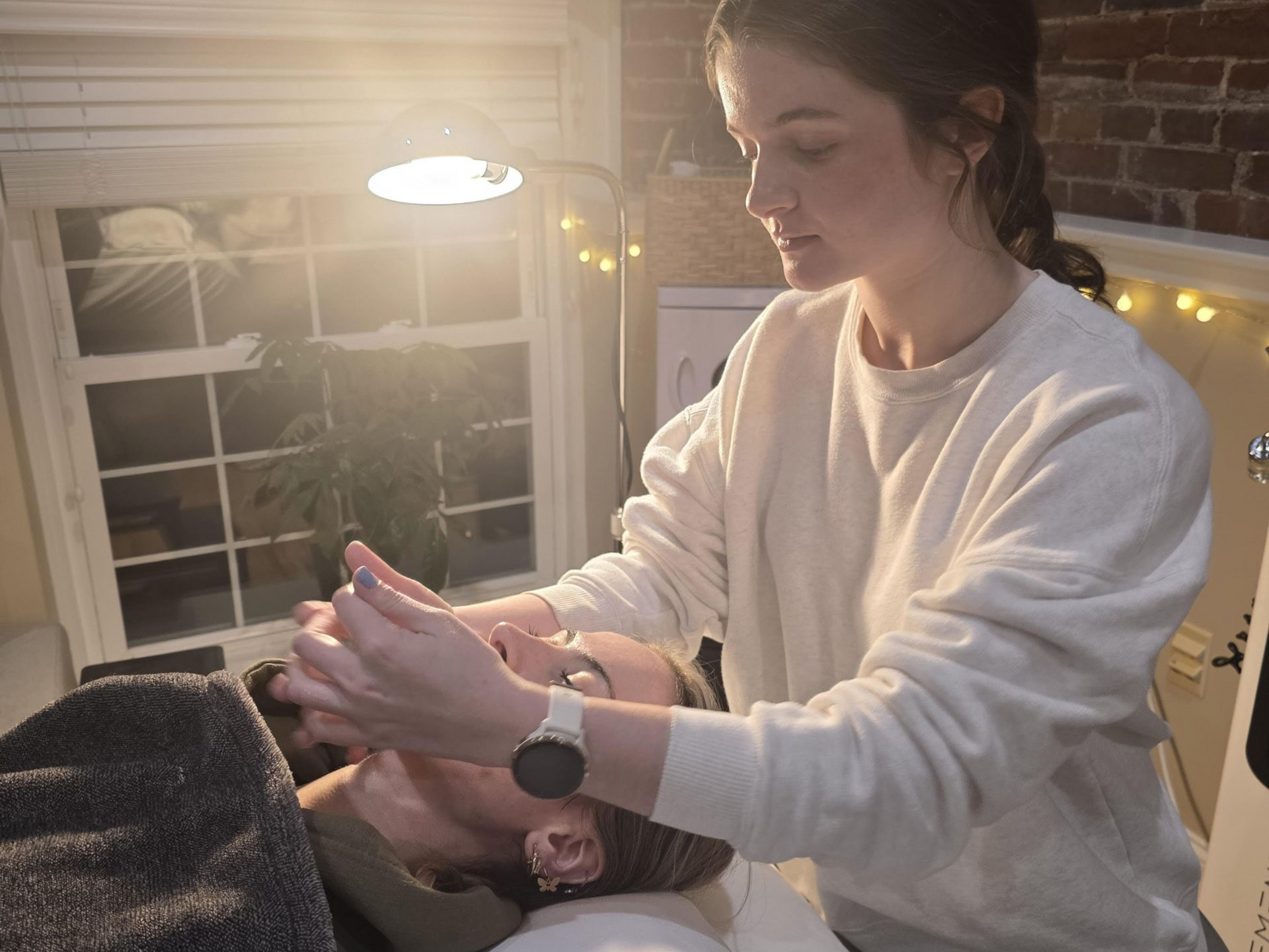 A person receiving a facial treatment in a room with a brick wall. Another person is performing the treatment.