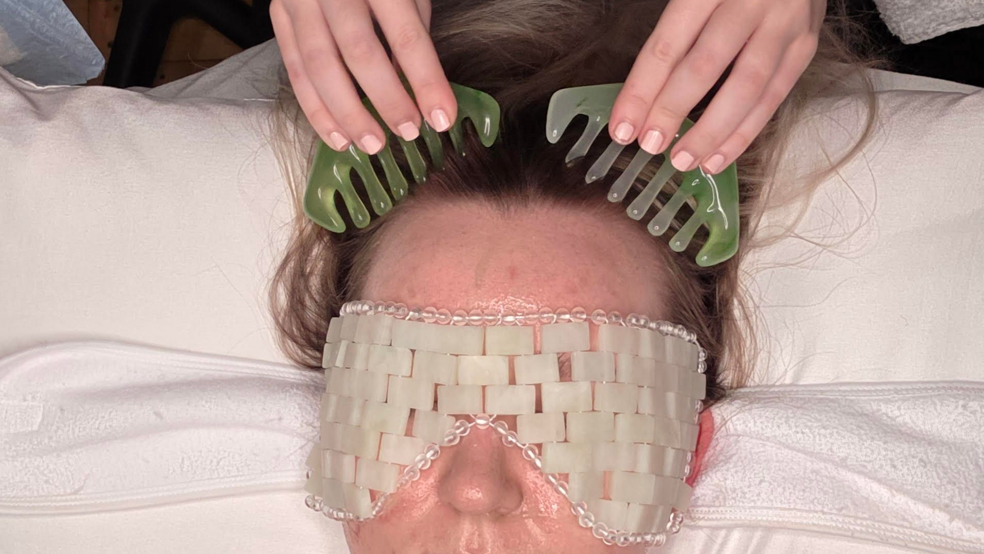 Person receiving a jade comb scalp massage and jade eye mask, on a white towel.
