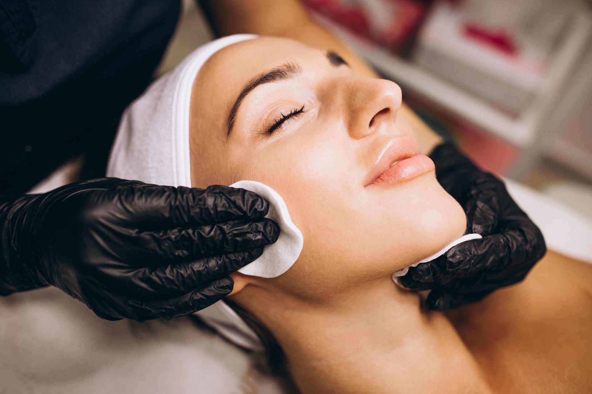 Gloved hands wiping a woman’s cheek with a cotton pad during a facial treatment