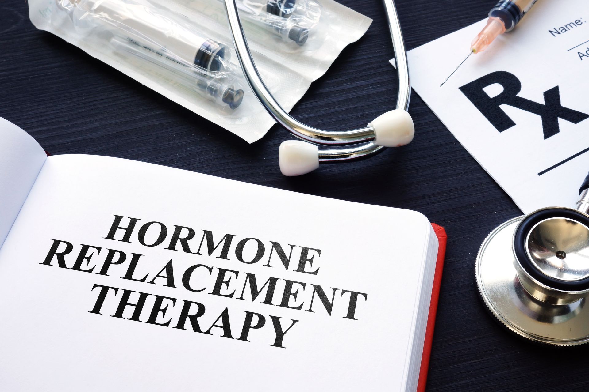 Hormone Replacement Therapy: book, stethoscope, syringes, and prescription on a dark surface.