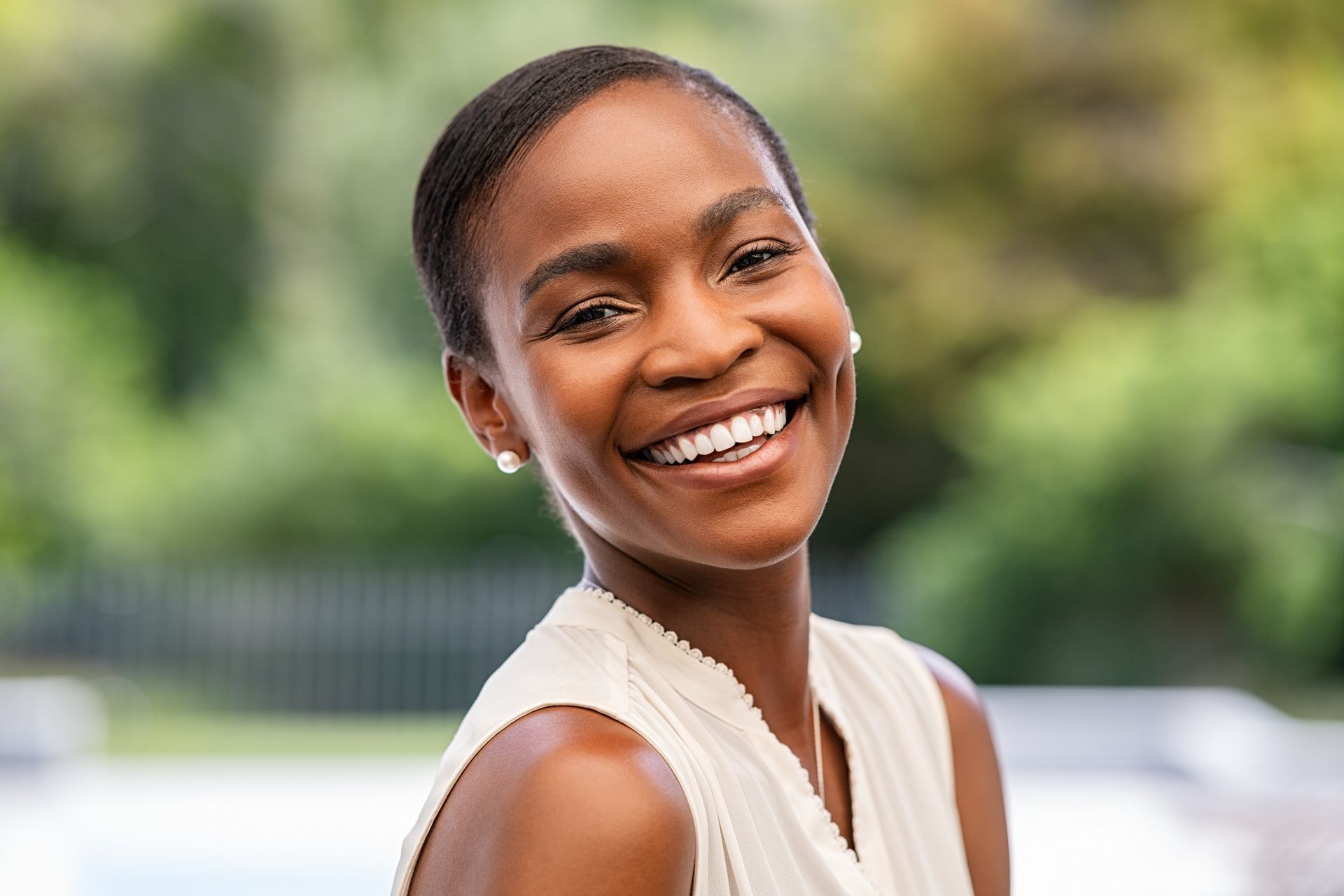 Smiling woman with short hair, wearing a white top, and pearl earrings, outdoors with blurred greenery in background.