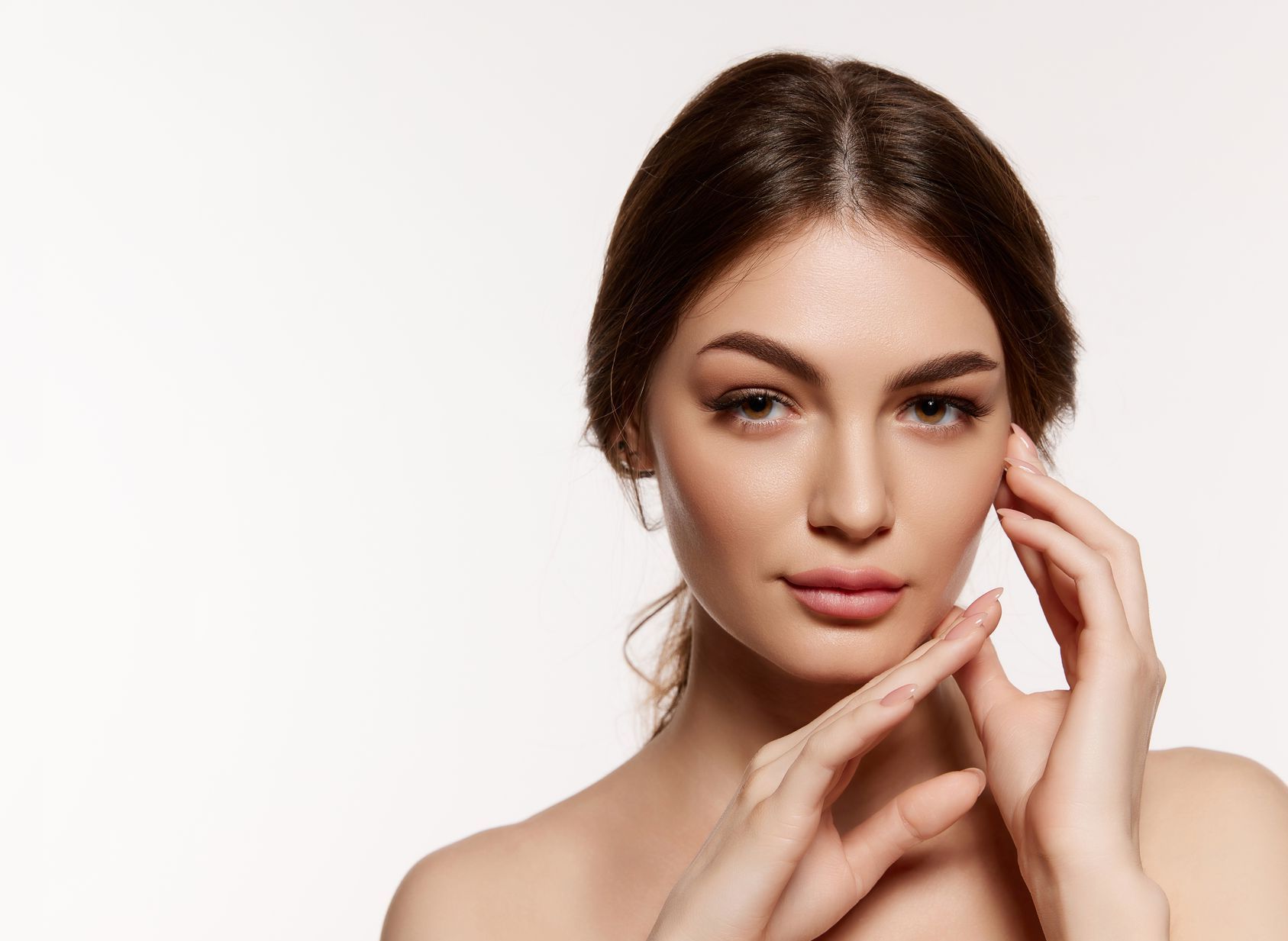 Woman with fair skin gently touches her face, looking towards the viewer, against a white backdrop.