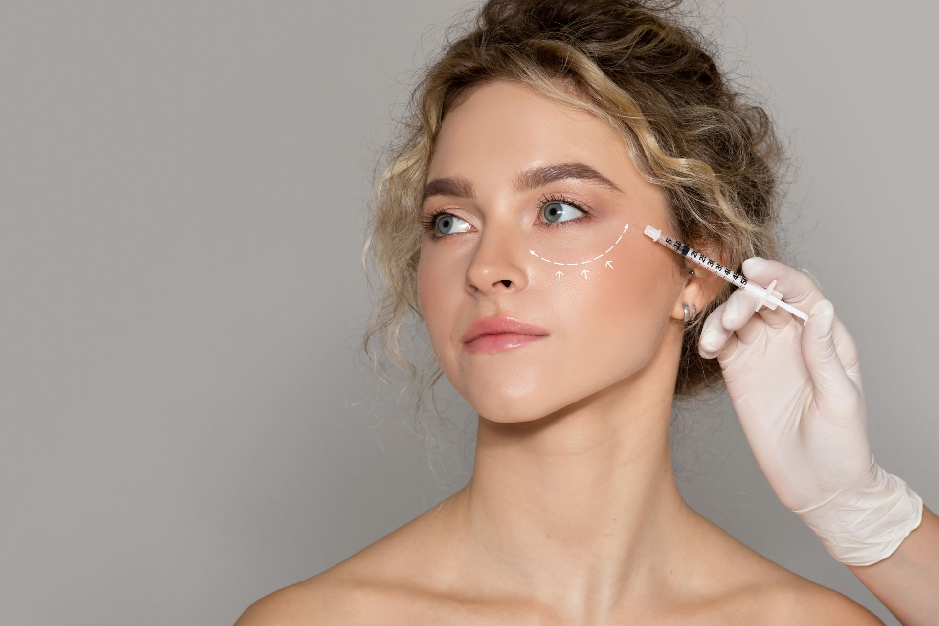 Gloved hand applying facial serum with a dropper to a woman’s cheek on a gray background
