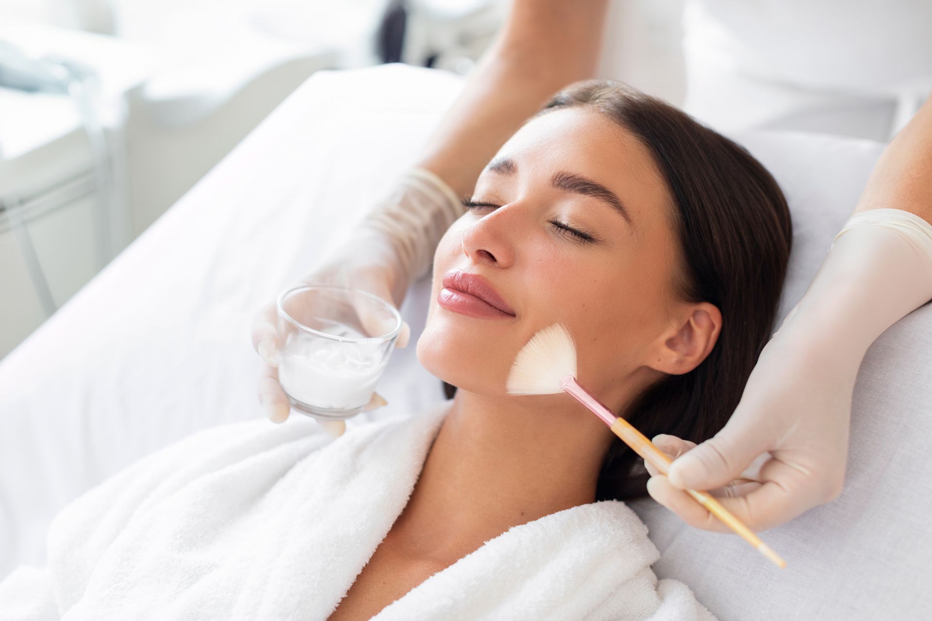 Woman receiving facial peel treatment, lying down with eyes closed. Hands in gloves, applying chemical substance.