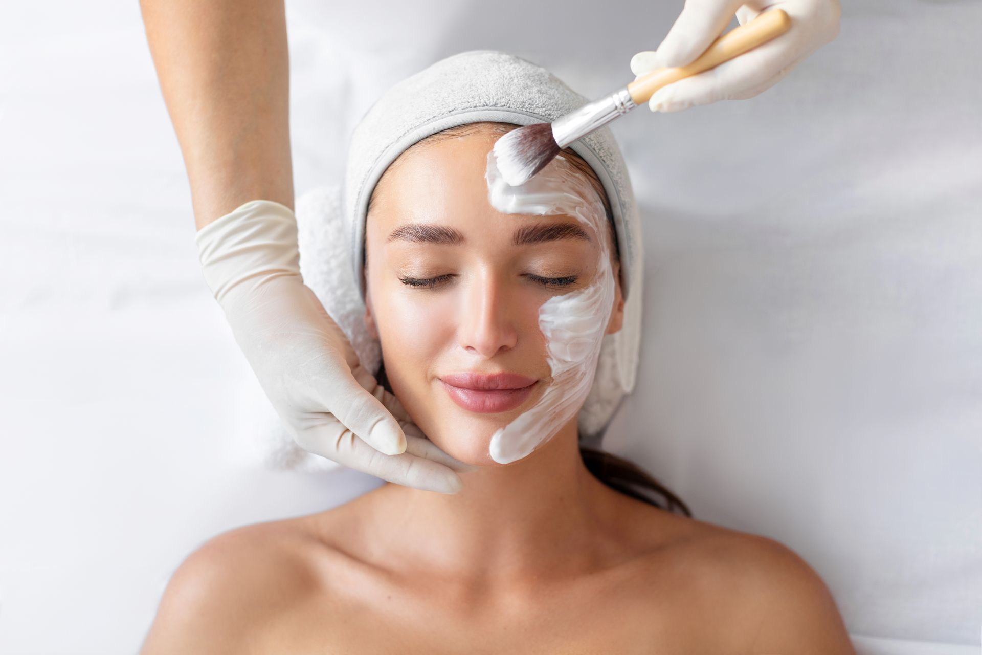 Facial treatment in progress as gloved hands apply cream to a woman’s face with a brush