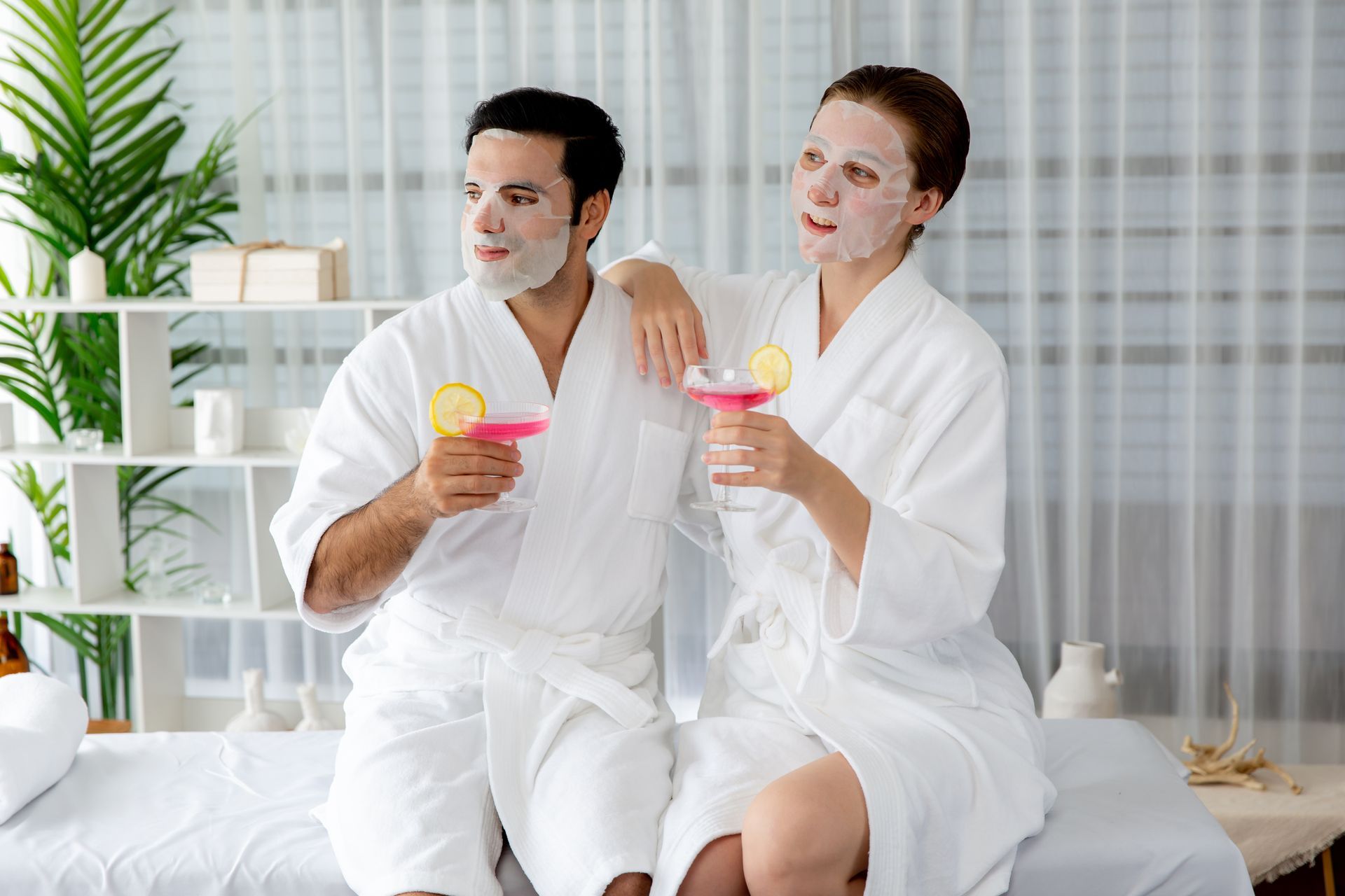 Couple in white robes with face masks holding pink drinks, relaxing in a spa-like setting.