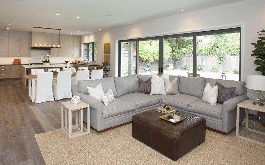 Bright modern living room with gray sectional, white dining set, and large windows overlooking greenery