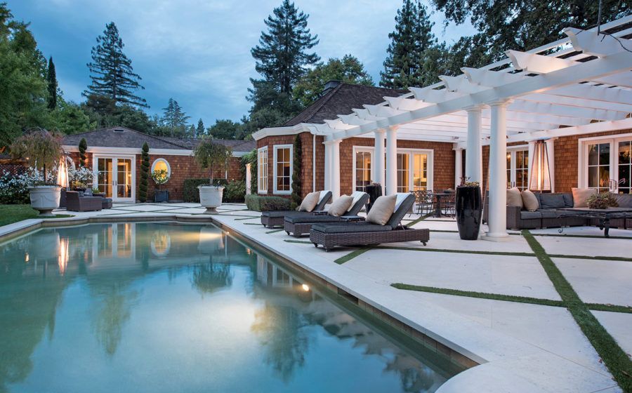 Luxury villa pool at dusk with lounge chairs, lit windows, and white pergola beside a reflective swimming pool