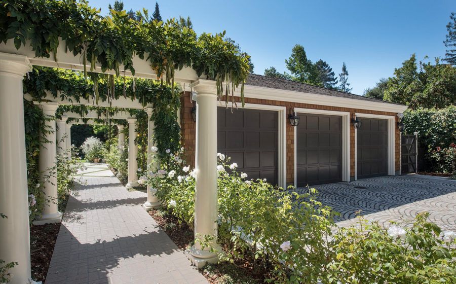 Sunlit courtyard with vine-covered archway, gravel path, and garage doors beside a stucco house