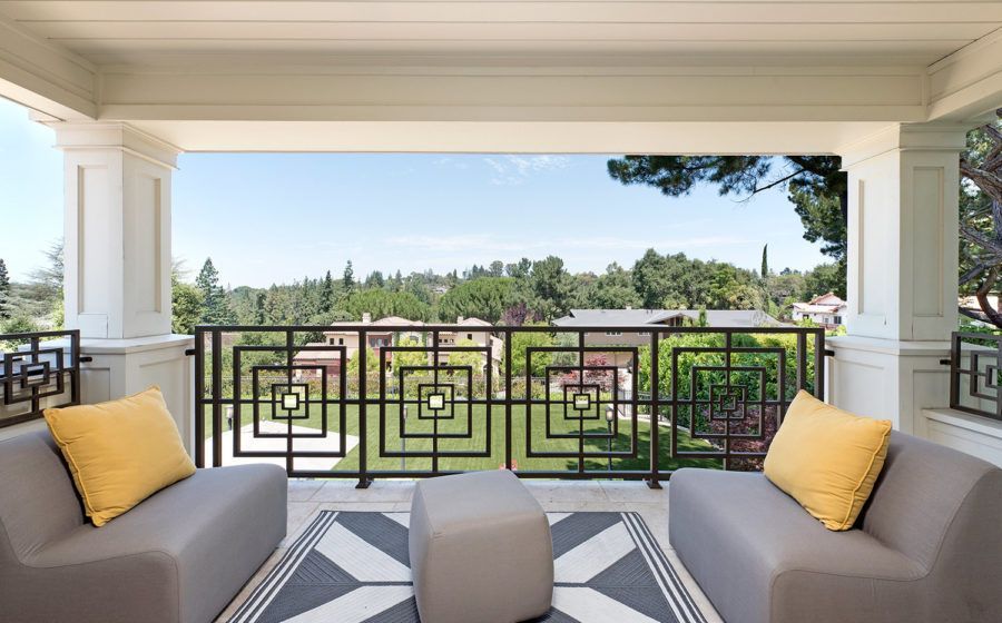 Covered patio with gray seating, yellow pillows, and a decorative railing overlooking a green landscape.