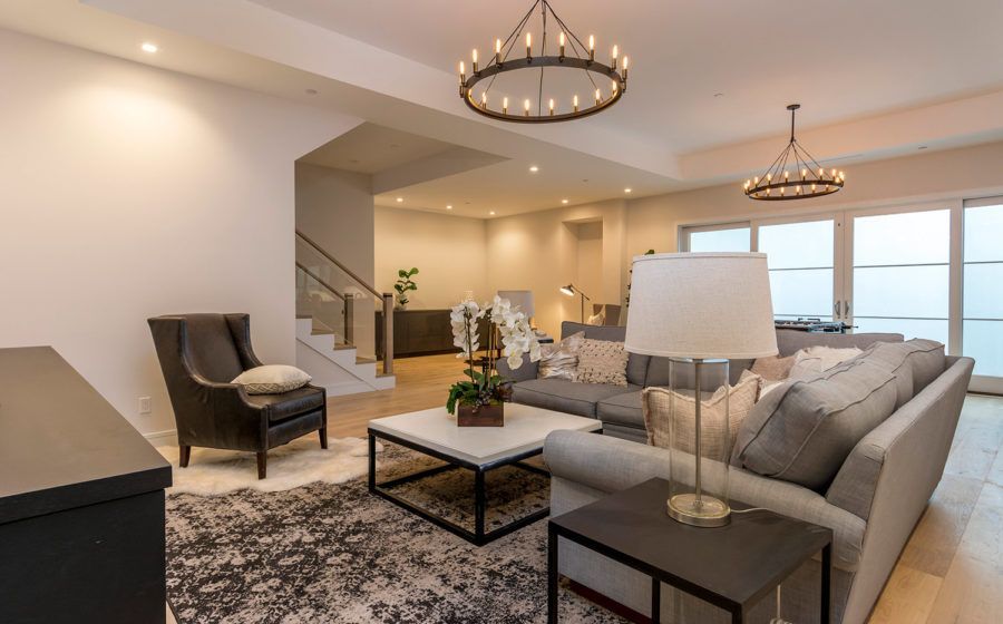 Modern living room with gray sectional, chandelier lighting, patterned rug, and large windows overlooking water