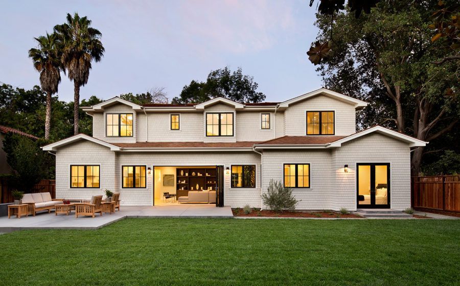 A large two-story white house with warm lit windows, a front porch, and a green lawn at dusk