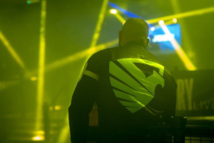 A man in a black jacket with a yellow eagle on the back is standing in a dark room.