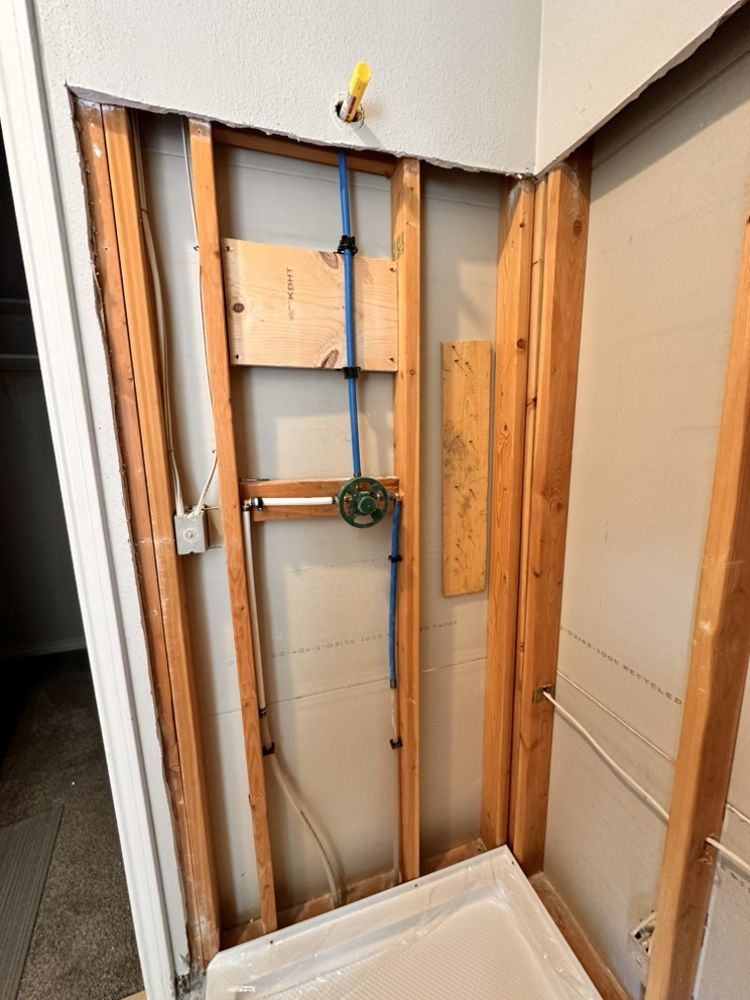 Framed shower plumbing: blue pipes, green valve, wood studs, white shower base, and pale walls.