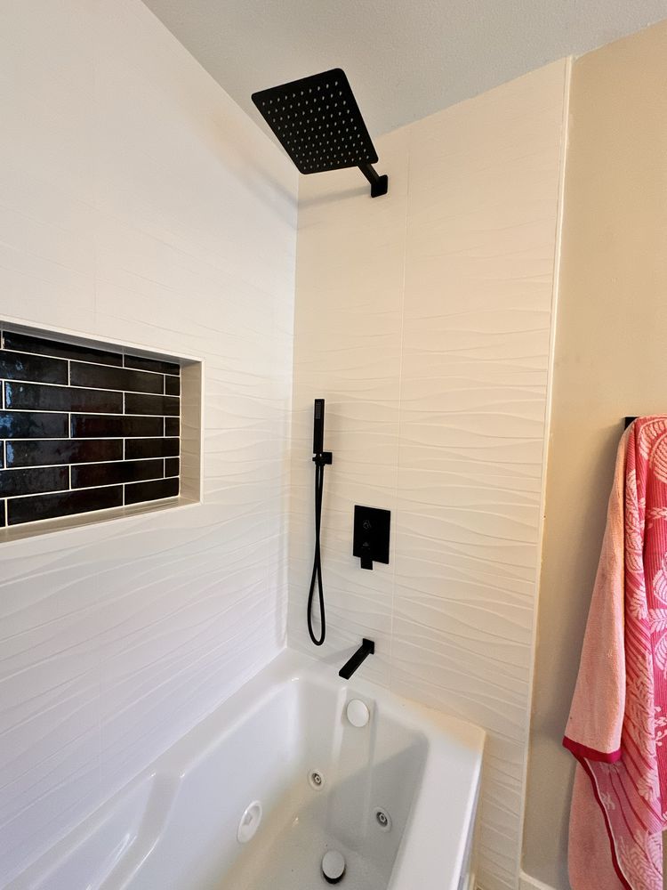 Black shower fixtures in a white tiled bathroom, including a rain shower head, with a black tile shelf.