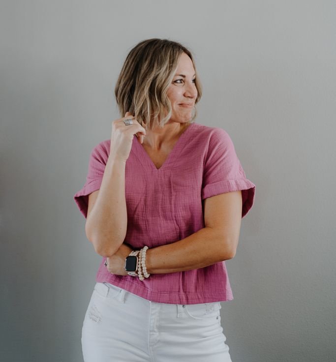 A woman wearing a pink top and white pants is standing in front of a wall.