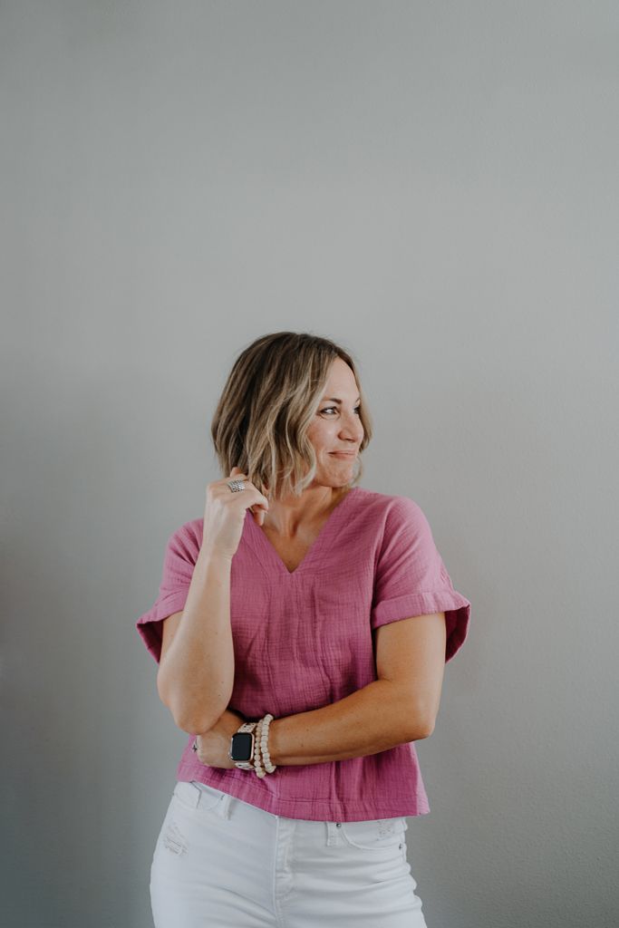 A woman in a pink top and white pants is standing in front of a wall.