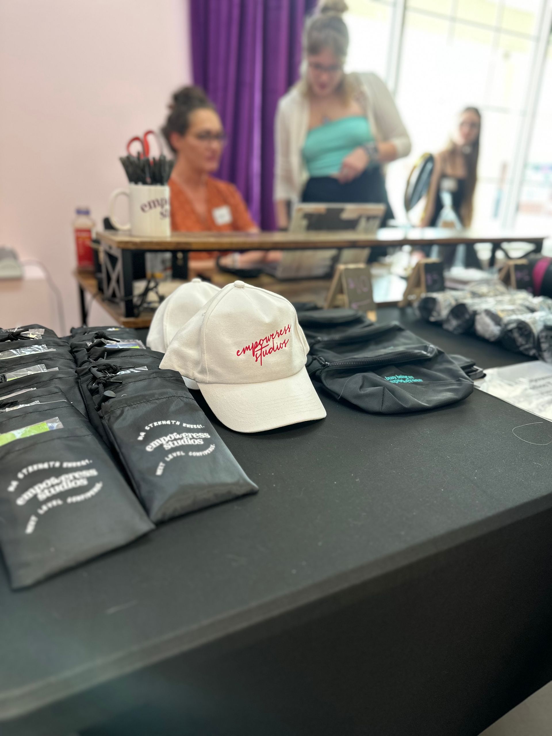 Merchandise on a black table; two women behind a desk in a shop. A white cap is featured.