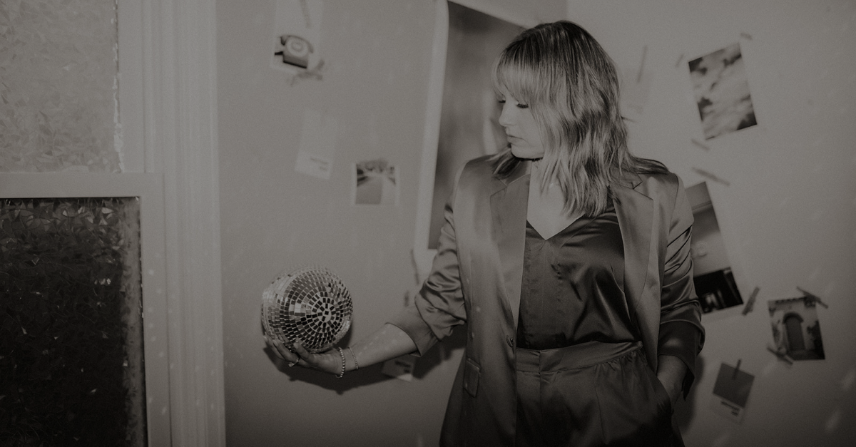 Woman in a satin outfit holding a small disco ball in a room with photos tacked to the wall.