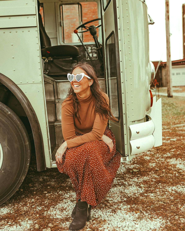 A woman wearing sunglasses is kneeling in front of a bus.