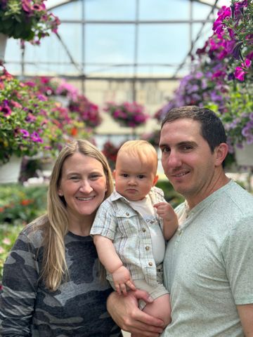 A man and woman are holding a baby in a greenhouse.