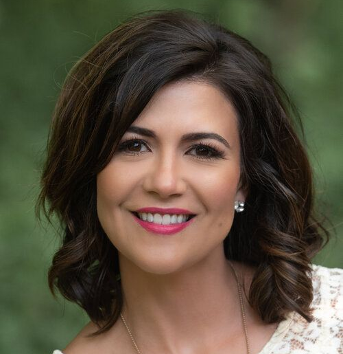 Woman with dark hair smiles, wearing a white lace top, outdoors, blurred green background.