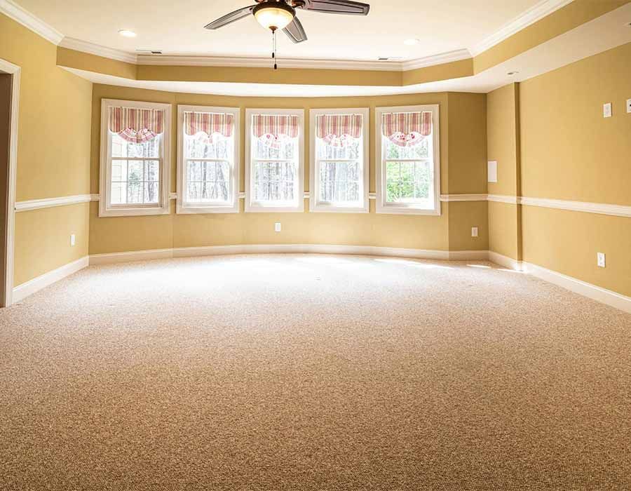 Empty beige carpeted living room with large windows and a ceiling fan