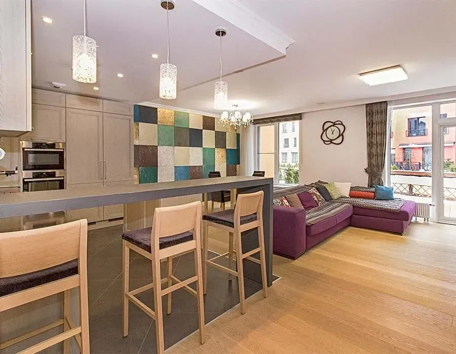 Modern open-plan kitchen and living room with bar stools, purple sofa, wood floors, and colorful tiled backsplash.