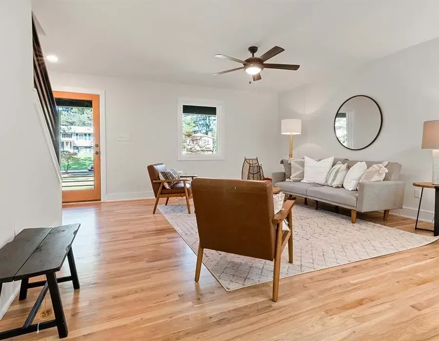 Bright living room with hardwood floors, gray sofa, tan chairs, round mirror, and ceiling fan