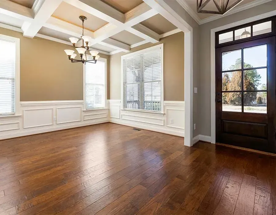 Empty living room with hardwood floors, beige walls, white wainscoting, chandelier, and large windows